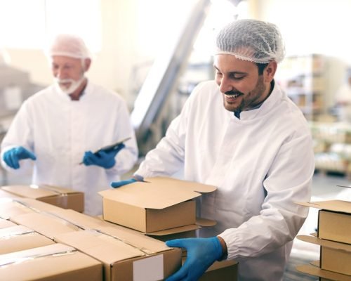 Young Caucasian man relocating boxes with cookies in food factory. In background his boss counting boxes and holding tablet.